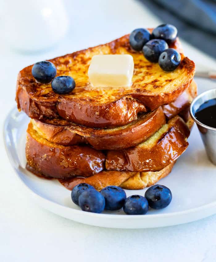 A stack of golden french toast on a white plate with blueberries and butter on top.