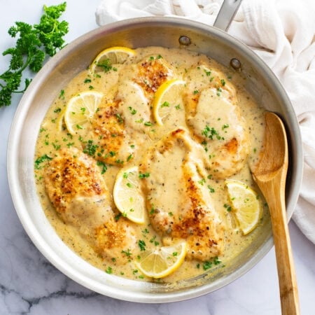 A skillet of Lemon Garlic Chicken with sauce, parsley, and lemons with a spoon on the side.