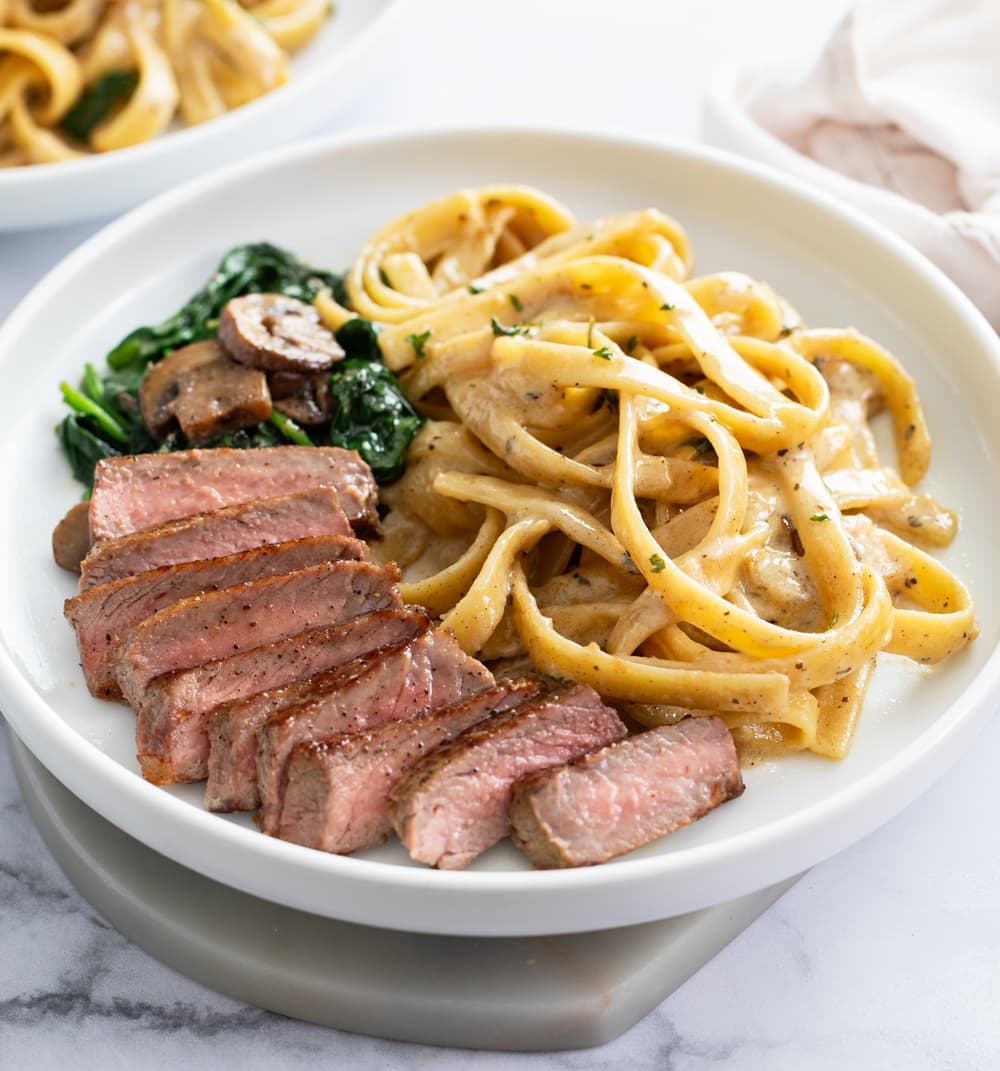 Steak pasta on a white plate with a creamy sauce, spinach, and mushrooms.